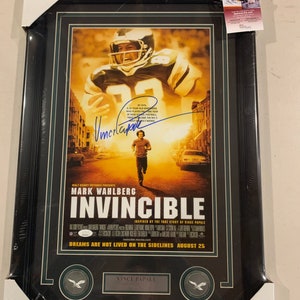 Op de afbeelding: Ingelijste filmposter voor de film "Invincible" met Mark Wahlberg. De poster toont een American footballspeler en de tekst "Invincible" in grote letters. De lijst is zwart met een groene rand en bevat een JSA-authenticatiesticker.
