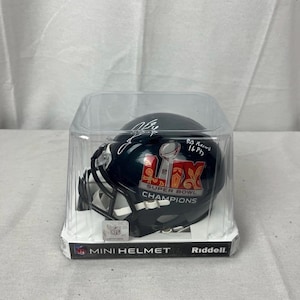 Puede incluir: Un mini casco de fútbol americano negro en una caja de plástico transparente. El casco tiene las palabras "SUPER BOWL CHAMPIONS" y el número romano "LIV" impresas. Está firmado y tiene el texto "EA Record 16 Pts". El embalaje dice "MINI HELMET" y "Riddell".