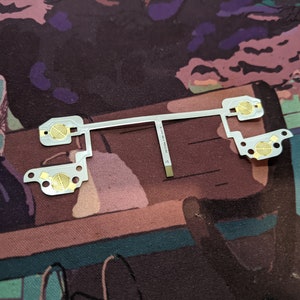 Op de afbeelding: Een wit printplaat met vier goudkleurige contactpunten. De printplaat is flexibel en heeft een dunne, zilveren draad die door het midden loopt. De tekst "W-1208-B017-V001-J1" is op de draad gedrukt.