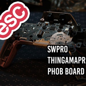 May include: A disassembled game controller housing featuring a black circuit board labeled Thingama Pro 2.1, with the text SWPRO THINGAMAPRO PHOB BOARD printed on the right and a red esc logo in the upper left corner, showing internal electronic components.