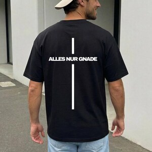 Könnte beinhalten: Ein schwarzes T-Shirt mit kurzen Ärmeln und einer großen weißen vertikalen Kreuzgrafik auf der Rückseite. Der Text ALLES NUR GNADE ist in weißen Großbuchstaben über den horizontalen Balken des Kreuzes gedruckt. Dazu eine hellblaue Jeans.