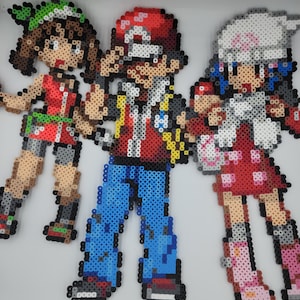 Puede incluir: Tres figuras de entrenadores Pokémon de pixel art. Las figuras están hechas de cuentas de colores. Las figuras están de pie sobre un fondo blanco. Las figuras son de una entrenadora con cabello verde y una camisa roja, un entrenador con cabello rojo y una camisa roja y una entrenadora con cabello blanco y una camisa roja.