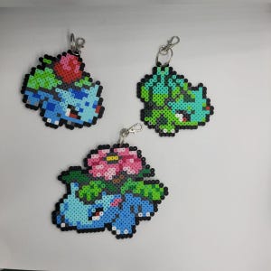 Peut inclure: Trois porte-clés en pixel art représentant Bulbizarre et ses évolutions. Les porte-clés sont faits de perles en plastique colorées, avec Bulbizarre en bleu, vert et rose. Chaque porte-clés a un fermoir en métal argenté.