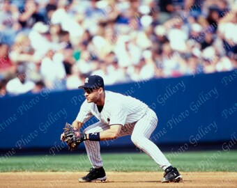 Puede incluir: Un jugador de béisbol con un uniforme a rayas blancas y azules, una gorra negra y gafas de sol está en posición de fildeo. El jugador lleva un guante negro y mira hacia el lado izquierdo de la imagen. El jugador está en un campo de béisbol con una valla azul y una multitud de personas en el fondo.
