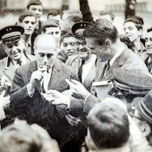 May include: A black and white photo of a group of people gathered around a man in a suit. The man is holding a cigarette and appears to be signing autographs. The photo is likely from the 1950s or 1960s.