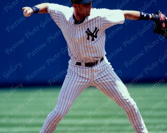 Puede incluir: Un jugador de béisbol con un uniforme a rayas blancas y azules con el logotipo de los New York Yankees en el pecho, está en posición de fildeo con un guante en su mano derecha. El jugador está en un campo de béisbol con un campo interior de hierba verde y un campo exterior de tierra marrón.