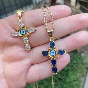 Op de afbeelding: Twee gouden kettingen met kruis hangers met blauwe en witte boze oog charmes. De kruis hangers zijn versierd met heldere en blauwe edelstenen.
