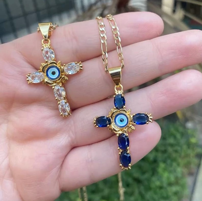 May include: Two gold necklaces with cross pendants featuring blue and white evil eye charms. The cross pendants are adorned with clear and blue gemstones.