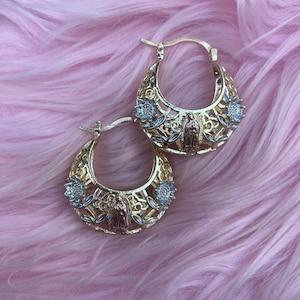 May include: Gold-tone hoop earrings with intricate filigree and floral details. Each earring features a central figure and accents of silver and rose gold. The earrings are set against a soft pink background.