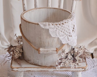 May include: A white wooden chair with a white wooden bucket on the seat. The bucket is decorated with a white crocheted doily and a rope handle. The chair is sitting on a white crocheted doily.
