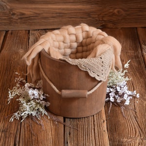 May include: A wooden bucket with a rope handle, lined with a beige knitted blanket and a crocheted lace trim. The bucket is sitting on a rustic wooden surface with white flowers.
