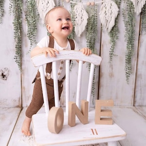 May include: A baby boy in brown pants and a white shirt stands on a white wooden chair. The chair has the word "ONE" spelled out in brown letters on the seat. The background is a white wood panel with a green garland.