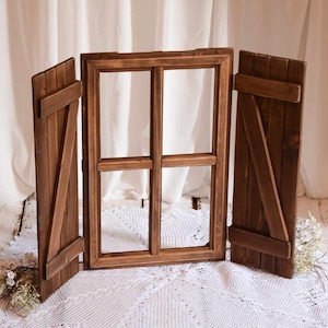 Op de afbeelding: Een houten raamkozijn met aan weerszijden twee luiken. De luiken zijn gesloten en het raamkozijn is leeg. Het raam staat op een wit oppervlak met een witte kanttafelkleed ervoor.