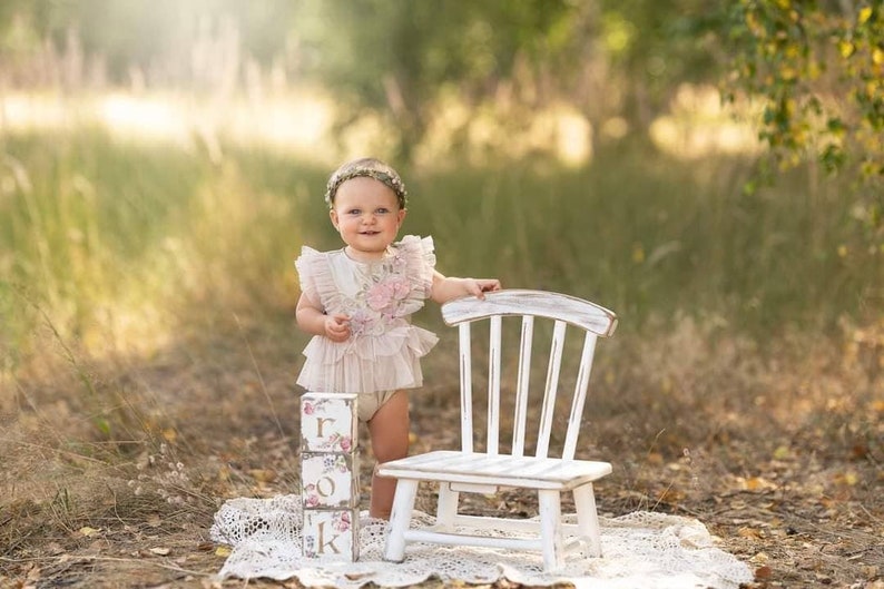 May include: A young child wearing a floral dress and a flower crown stands in a grassy field. The child is holding a white wooden chair and a stack of wooden blocks that spell "ONE".