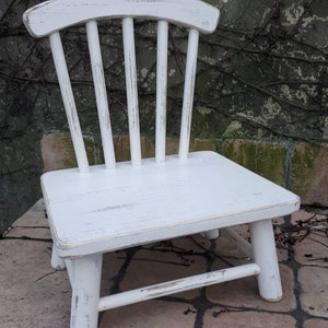 May include: White wooden chair with a distressed finish. The chair has a simple design with a curved back and four legs. The chair is sitting on a stone patio.