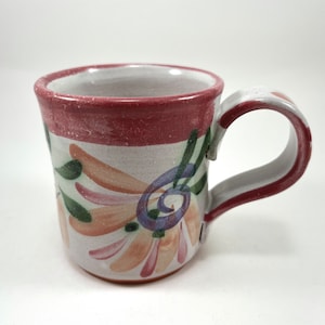 May include: A white ceramic mug with a red rim and handle. The mug is decorated with a floral design in shades of pink, green, and purple.