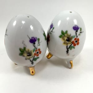 May include: A pair of white ceramic salt and pepper shakers shaped like eggs with gold feet. The shakers are decorated with colorful floral designs.