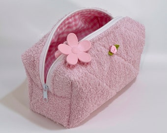 May include: A pink quilted cosmetic bag with a white zipper and a pink flower hair clip. The interior lining features a pink and white gingham pattern. A small pink rose is attached to the exterior.