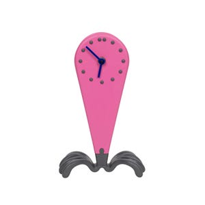 Puede incluir: Un reloj rosa con una aguja de hora y minutos azules. El reloj tiene un diseño único con una parte superior puntiaguda y una base con tres patas curvas.