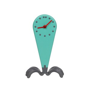 Op de afbeelding: Een tealblauwe klok met een rode uurwijzer en een zwarte minutenwijzer. De klok heeft een uniek ontwerp met een puntige bovenkant en een voet met drie gebogen poten.