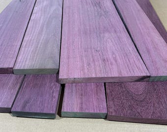 May include: A collection of purpleheart wood planks, showcasing a range of violet hues. The planks are arranged in a layered display, revealing the natural grain and texture of the wood. The wood appears to be freshly cut and ready for use.