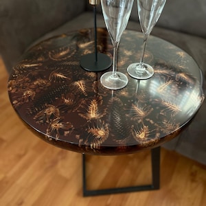 May include: A round, dark brown side table with a black metal base. The table top has a unique design featuring a pattern of light brown pine cones embedded in resin.