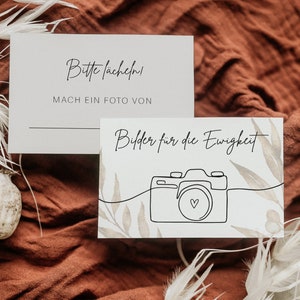 May include: Two light grey cards with black script on a rust-coloured fabric background. One card reads "Bitte lächeln! MACH EIN FOTO VON" and the other card has a line drawing of a camera with the text "Bilder für die Ewigkeit".