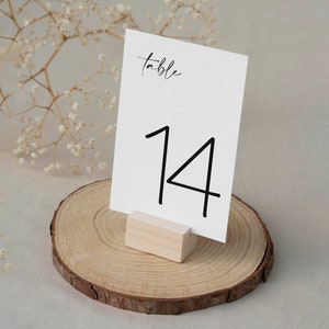 Könnte beinhalten: Eine weiße Tischkarten mit der schwarzen Zahl 14, präsentiert auf einem Holzblock und einer Holzscheibenbasis. Das Wort "table" ist in einer zarten Schrift oben auf der Karte geschrieben. Der Hintergrund ist neutral und hell.