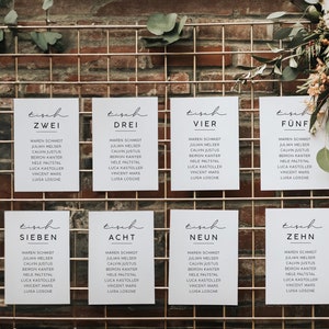 May include: A display of ten white cards with black text, arranged on a gold grid. Each card has the word "Tisch" and a number, followed by names. The background is a brick wall with floral arrangements.