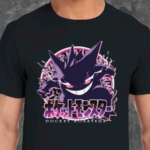 Può includere: T-shirt nera con grafica viola e bianca di un personaggio dei cartoni animati e testo giapponese. Il design presenta un volto sorridente e punte. Il testo recita "Pocket Monsters". La maglietta è realizzata in materiale morbido.