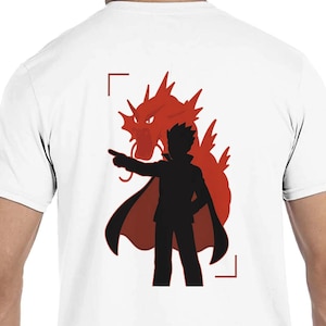 May include: White t-shirt featuring a black silhouette of a person with a red cape, pointing towards a red dragon. The design is framed by red corner brackets.