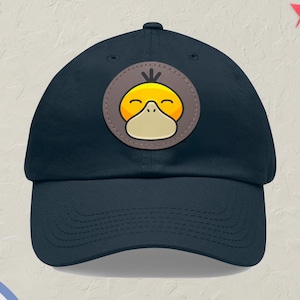 May include: Navy blue baseball cap with a circular patch featuring a yellow cartoon duck face with a beige beak and a gray background. The cap has a curved brim and a top button.