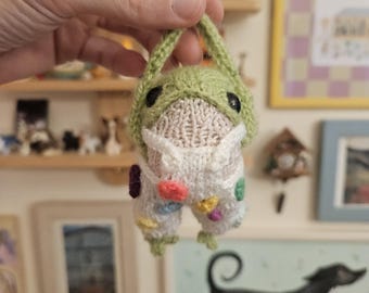 May include: A small, hand-knitted frog figurine featuring a light green hood and a white body. The frog is wearing white overalls decorated with small, colorful, circular patches in shades of yellow, pink, purple, and green. It has two black eyes.