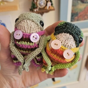 May include: Two small, round, knitted creatures with black bead eyes and two light purple buttons on their chests. The left creature has green, brown, and purple stripes, while the right one features green, yellow, and orange stripes with a green cap.
