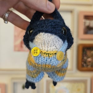 May include: A small, handmade knitted plush toy featuring a dark navy blue head with black plastic eyes and a white chin. The creature wears light blue and yellow striped knitted overalls secured with two small, round, bright yellow buttons.