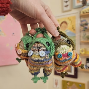 May include: Three handmade, colorful knitted frog ornaments. Each frog has button eyes, a green hat, and striped pants. The frogs are held up by a hand, showcasing their unique, whimsical design.