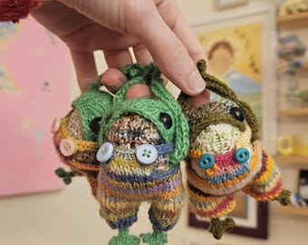 May include: Three handmade, colorful knitted frog ornaments. Each frog has button eyes, a green hat, and striped pants. The frogs are held up by a hand, showcasing their unique, whimsical design.