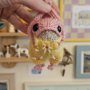 May include: A small, handmade crochet creature featuring a fuzzy yellow body, a light gray face, and a pink knitted bonnet with two black bead eyes. A decorative bow made of small, multicolored glass beads is attached to the front of the body.