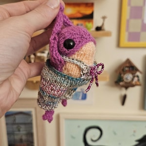 May include: A small, handcrafted crochet creature featuring a vibrant purple head with a large black eye, wearing a multi-colored, speckled knit sweater. The figure is adorned with a delicate, thin beaded string tied into a bow around its neck.