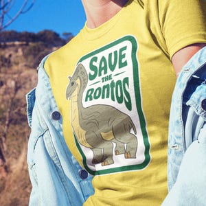 Op de afbeelding: Geel T-shirt met een groene en witte afbeelding van een dinosaurus met de tekst "Save the Ronto's" erop.