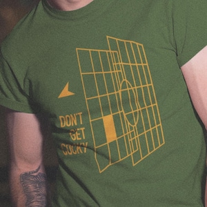 Puede incluir: Una camiseta de manga corta color verde oliva con un estampado gráfico amarillo de una estructura de rejilla que recuerda a una pantalla de ordenador de puntería. El diseño incluye un icono de flecha y el texto "DON'T GET COCKY" en amarillo.