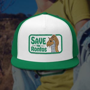 Op de afbeelding: Groene en witte truckerpet met de tekst "SAVE the Ronfos" en een cartoonillustratie van een Ronfos. De pet heeft een groene klep en mesh achterkant, met een wit voorpaneel.