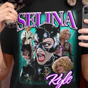 Pode incluir: Camiseta preta com um gráfico de Selina Kyle, o texto "Selina Kyle" em uma fonte retrô roxa e branca. O design inclui várias imagens da personagem em poses diferentes.