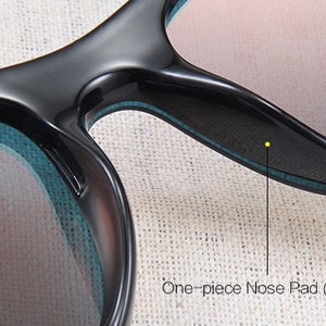 May include: Close-up of black sunglasses with a one-piece nose pad for comfort. The lenses have a gradient tint, and the frames have a sleek, modern design. The text "One-piece Nose Pad (more comfortable)" is visible.