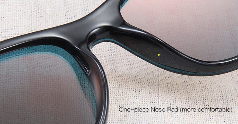May include: Close-up of black sunglasses with a one-piece nose pad for comfort. The lenses have a gradient tint, and the frames have a sleek, modern design. The text "One-piece Nose Pad (more comfortable)" is visible.
