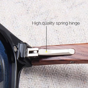 May include: Close-up of sunglasses with a black frame and blue lenses. The wooden arm features a high-quality spring hinge. The text "High quality spring hinge" is visible.