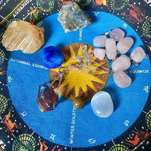 May include: A blue and black circular fabric with zodiac signs and symbols.  The fabric has a variety of crystals and stones arranged around the center, including a clear bottle with a silver chain, a light blue stone, a pink stone, a brown stone, a dark blue stone, a light brown stone, and a red stone.