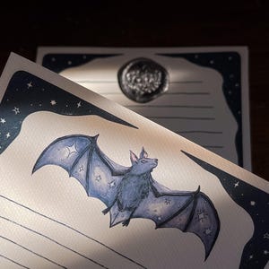 May include: A stationery set featuring a watercolor illustration of a bat with star accents. The bat is a muted blue color, and the stationery has a dark blue border with stars. A wax seal is visible in the background.