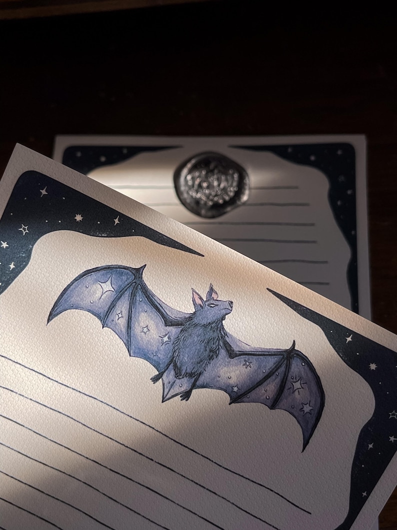 May include: A stationery set featuring a watercolor illustration of a bat with star accents. The bat is a muted blue color, and the stationery has a dark blue border with stars. A wax seal is visible in the background.