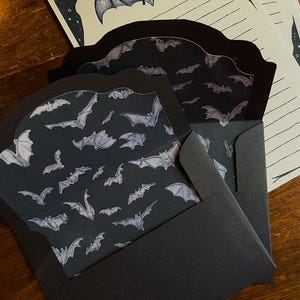 May include: Black stationery set featuring bat illustrations. The set includes envelopes with a bat pattern, lined paper, and a decorative bat illustration. The envelopes are black, and the paper has a starry border.